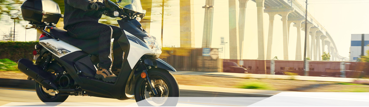 Person riding a black Yamaha Zuma motorcycle on a street under a white bridge.